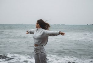 A woman stands with arms spread by the sea, embracing the freedom of a windy, overcast day.